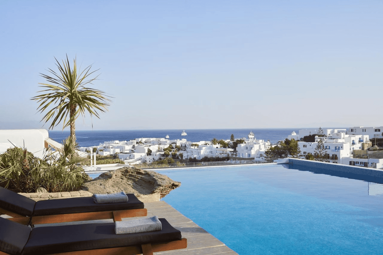 Pool overlooking Mykonos town and Aegean Sea, with sun loungers and scenic island views.