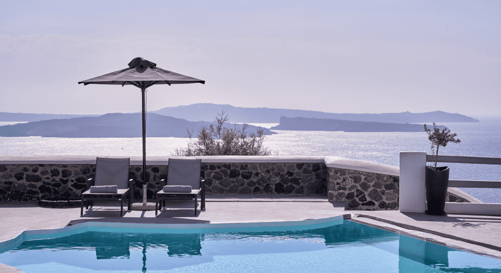 Scenic view of a pool with lounge chairs and umbrella overlooking the sea in Oia, Santorini.